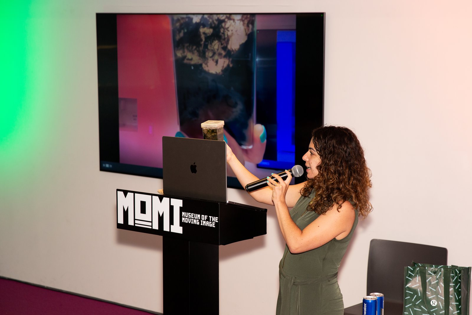 Ana Gonzalez-Nayeck speaking into a microphone at a Museum of the Moving Image podium, holding up a jar of microbial mat while a close-up of it plays on the screen behind her